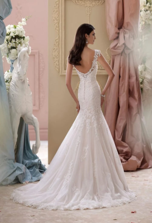 Emerson from David Tutera is a lace, cap sleeve wedding gown. A luxurious fit and flare wedding dress with a sweetheart neckline features an illusion and lace modesty piece, lace bodice with exaggerated dropped waistline, illusion  deep plunging open back  finished with  covered button closures, scalloped hemline and chapel length train